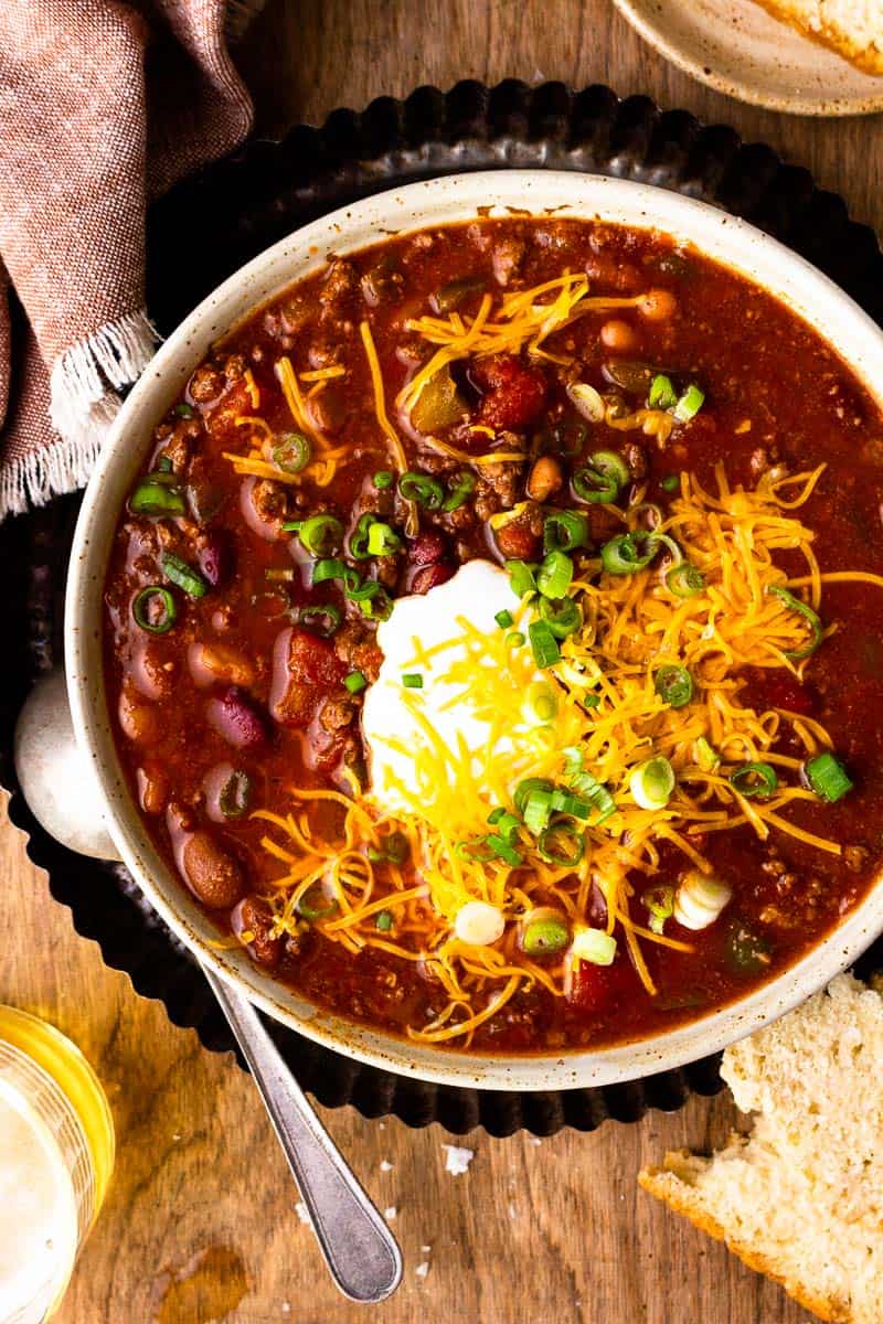 Venison chili in a bowl topped with shredded cheese and sour cream. Sitting next to a spoon, beer and slice of bread.