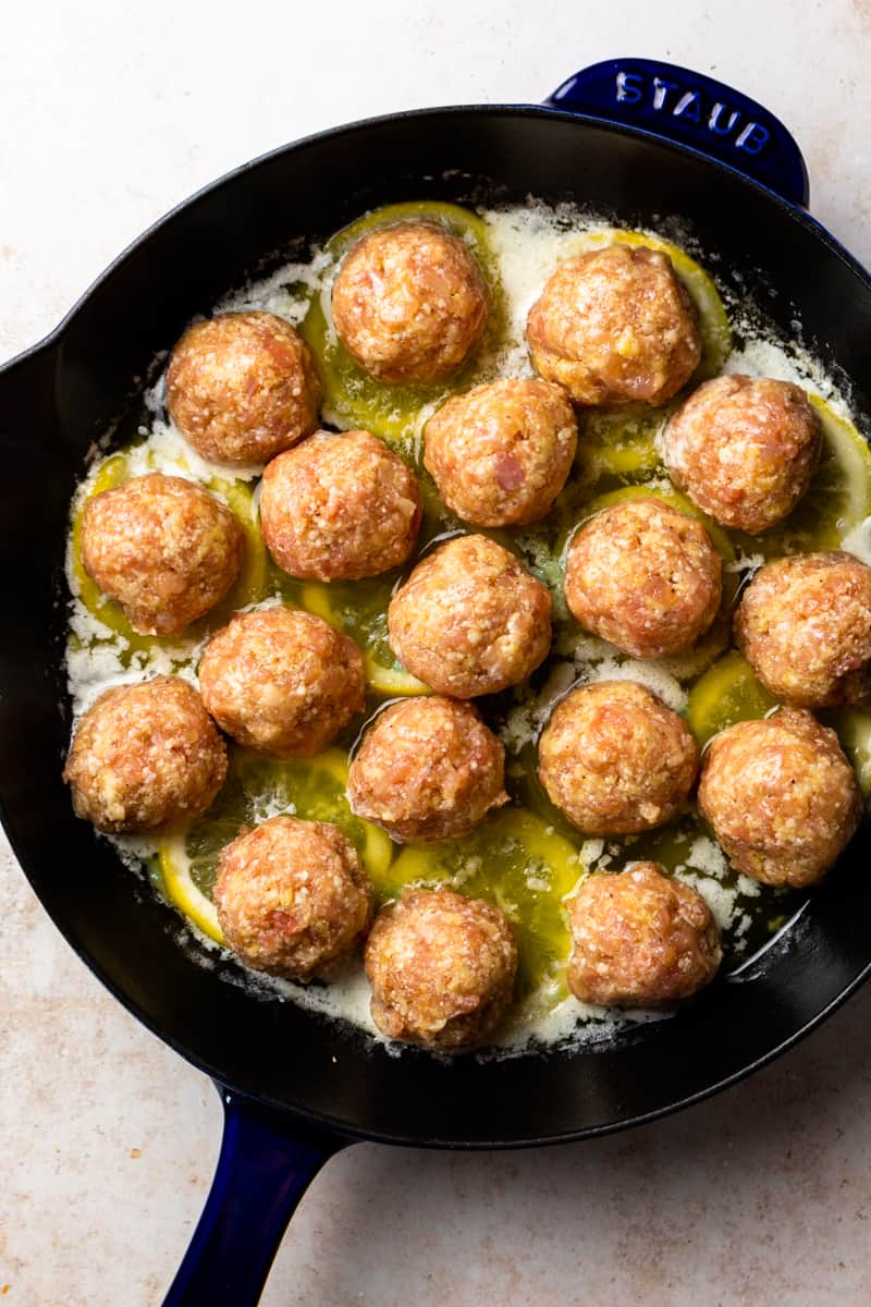 uncooked chicken meatballs in a skillet with melted butter, lemon slices and garlic