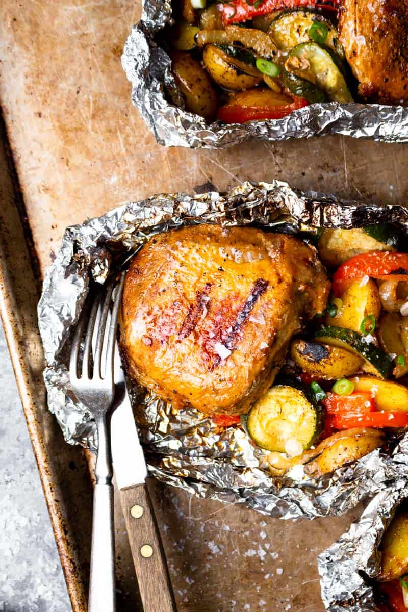 grilled pork chop and vegetables in an open foil pack with a fork and knife on a sheet pan