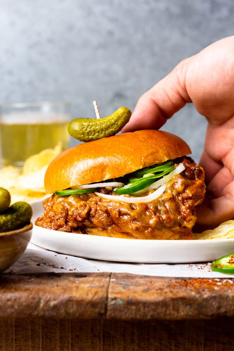 hand picking up a chili cheese sloppy joe sandwich served on a plate with potato chips