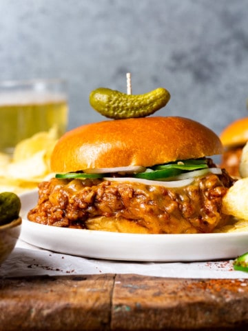 chili cheese sloppy joe meat between a hamburger bun served on a plate with potato chips