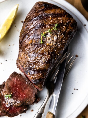 Sliced venison backstrap on a plate with a fork, knife and lemon wedge.