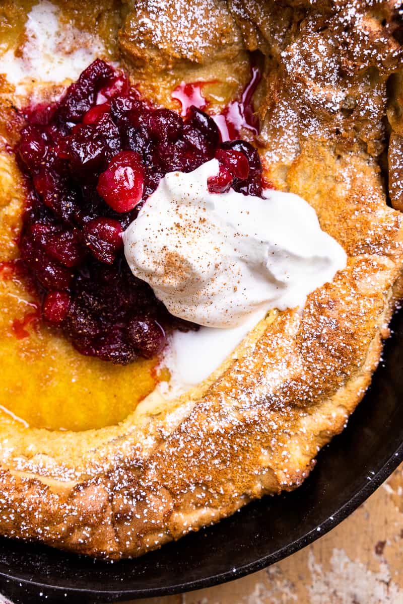 dutch baby in a skillet topped with powdered sugar, spiced cranberries, and whipped cream