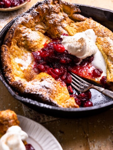 dutch baby in a skillet topped with powdered sugar, spiced cranberries, and whipped cream