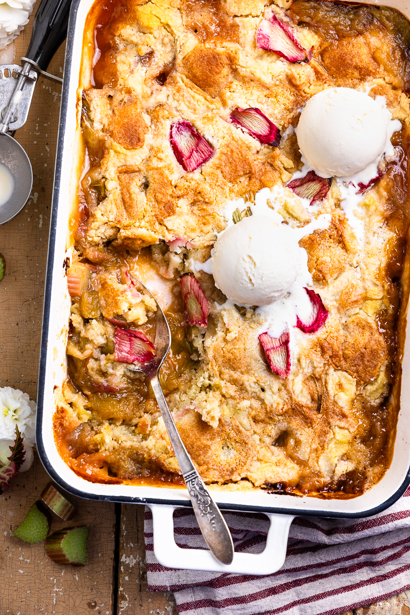 Rhubarb dump cake in a baking dish topped with vanilla ice cream and using a spoon to scoop out cake