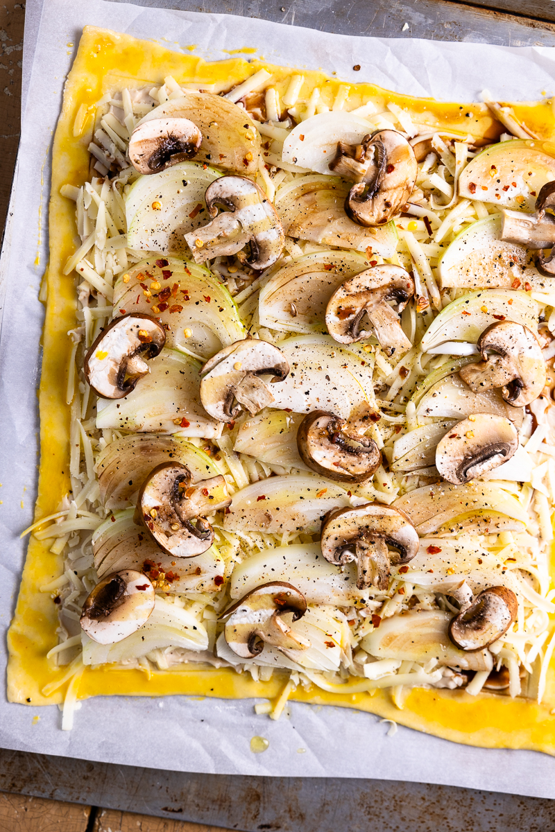 puff pastry spread with French onion dip and topped with white cheddar cheese, onions and mushrooms, then seasoned with red pepper flakes, salt and pepper with egg wash on the edges