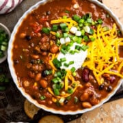 Bowl of venison chili topped with cheese and green onions sitting next to a cup of beer and crackers.