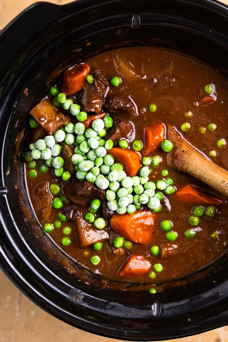 Cooked venison stew in a slow cooker with frozen peas on the top before mixing them in.