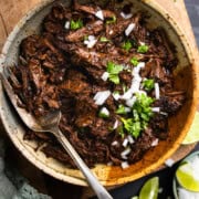 Shredded venison barbacoa in serving bowl next to a small bowl of diced onion, small bowl of chopped cilantro, lime wedges and a beer.