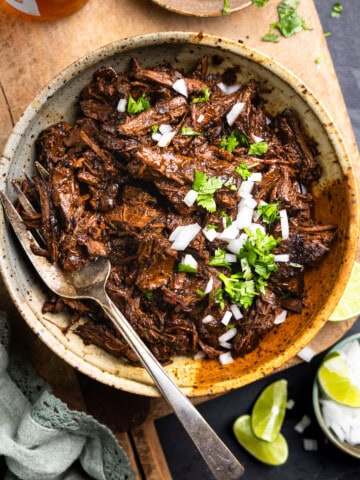 Tender venison barbacoa in a bowl topped with diced onion and cilantro, served with lime wedges and beer.