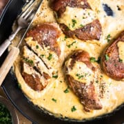 Pan-seared pheasant breasts in a skillet with dijon mustard cream sauce and garnished with fresh parsley sitting next to a glass of white wine, bowl of chopped parsley and a black pepper pinch bowl.