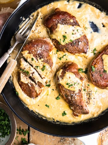 Pan-seared pheasant breasts in a skillet with dijon mustard cream sauce and garnished with fresh parsley sitting next to a glass of white wine, bowl of chopped parsley and a black pepper pinch bowl.