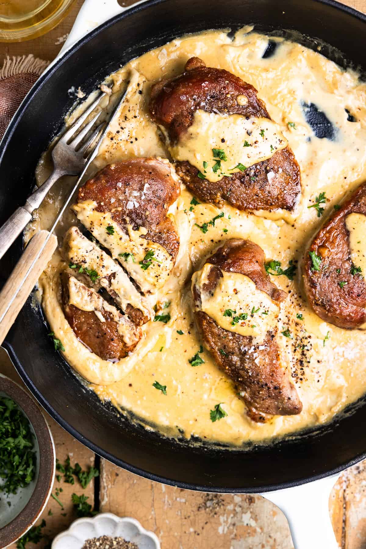 Pan-seared pheasant breasts in a skillet with dijon mustard cream sauce and garnished with fresh parsley sitting next to a glass of white wine, bowl of chopped parsley and a black pepper pinch bowl.