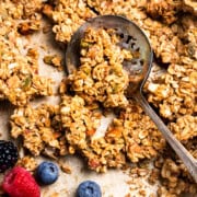 Scooping sourdough granola from a sheet pan next to a bowl of fresh berries.