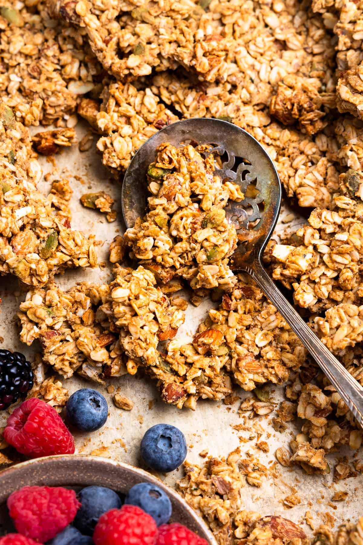 Scooping sourdough granola from a sheet pan next to a bowl of fresh berries.