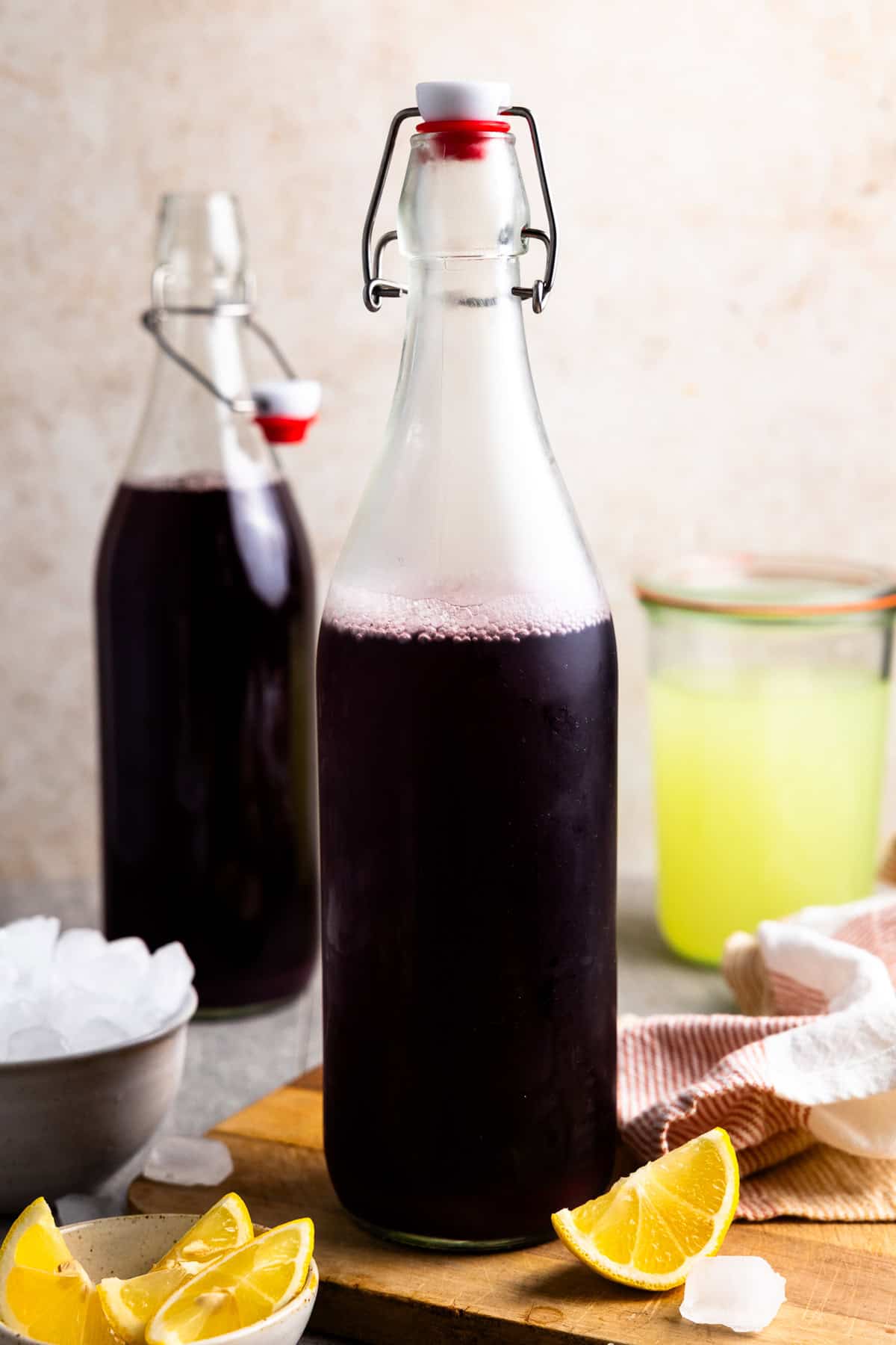 Grape whey soda in a swing top bottles siting next to a jar of whey, bowl of ice, and bowl of lemon wedges.