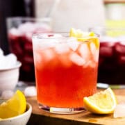 Cranberry pomegranate fermented whey soda in a glass with ice and a lemon wedge sitting next to two other cups of whey soda and bowl of lemon wedges.