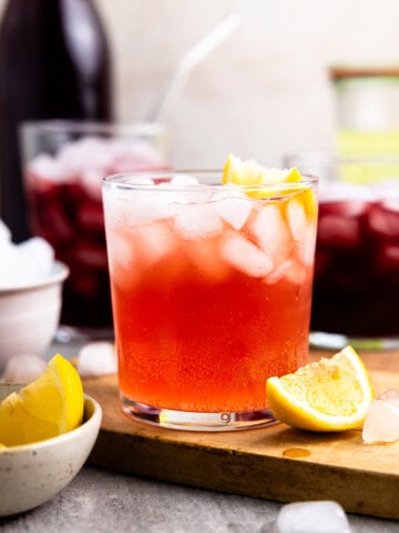 Cranberry pomegranate fermented whey soda in a glass with ice and a lemon wedge sitting next to two other cups of whey soda and bowl of lemon wedges.