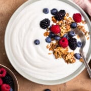 Yogurt in a bowl topped with granola and berries. Sitting next to a bowl of berries, bowl of granola and a cup of orange juice.
