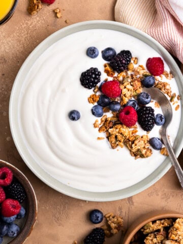 Yogurt in a bowl topped with granola and berries. Sitting next to a bowl of berries, bowl of granola and a cup of orange juice.