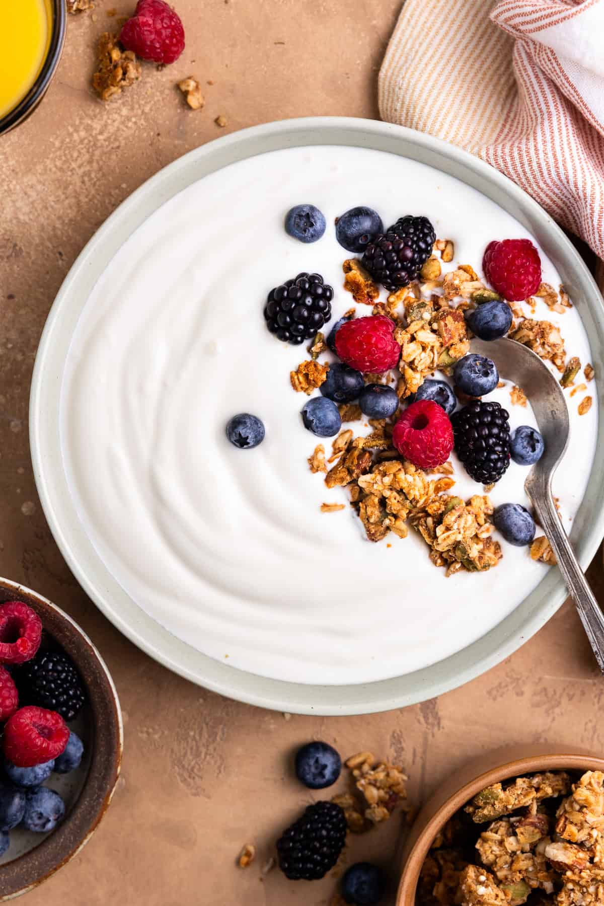 Yogurt in a bowl topped with granola and berries. Sitting next to a bowl of berries, bowl of granola and a cup of orange juice.