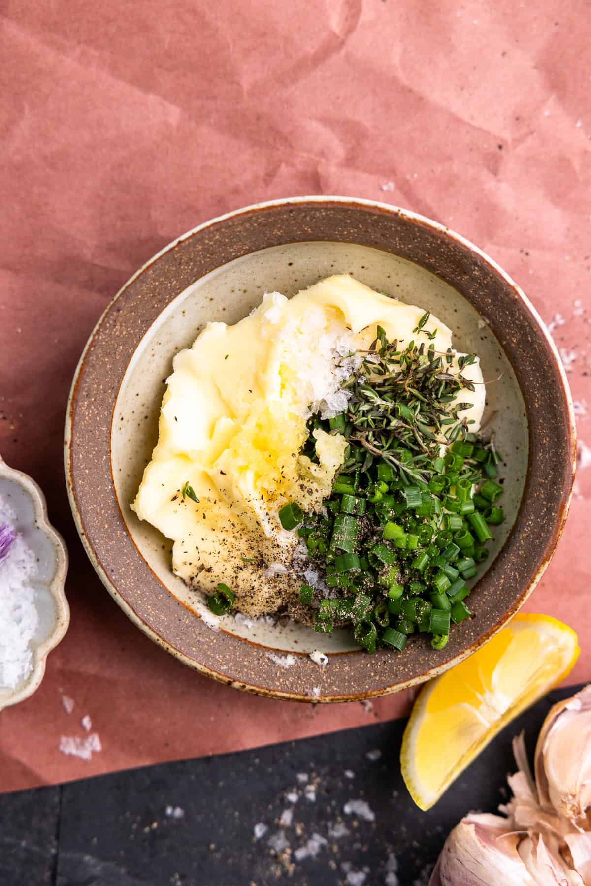 Butter in a bowl topped with salt, pepper, garlic, fresh thyme, and chives to make garlic herb butter for steak.