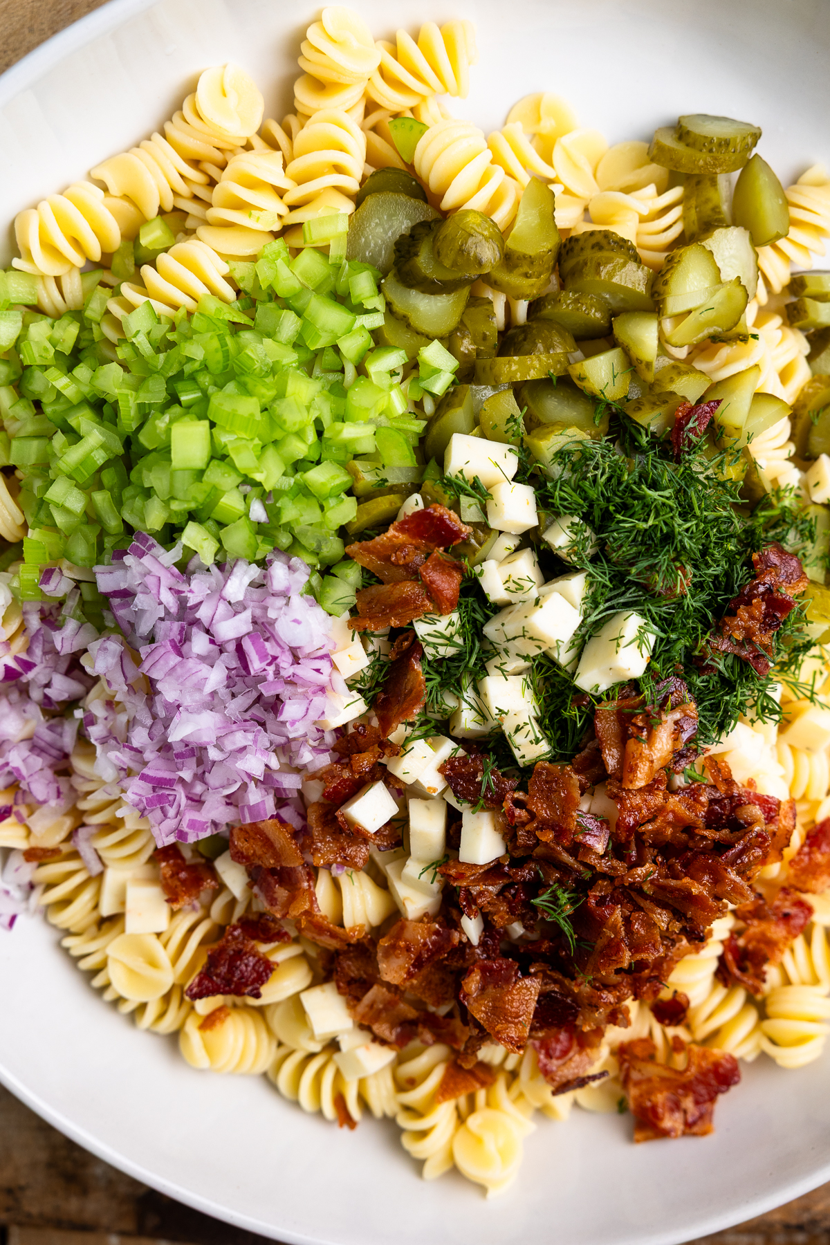 Pasta, bacon, celery, onion, dill, pickles, and cheese all in a large bowl.