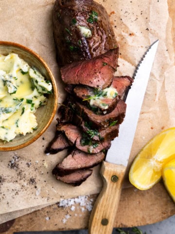 Venison tenderloin sliced on a cutting board and topped with herb butter.